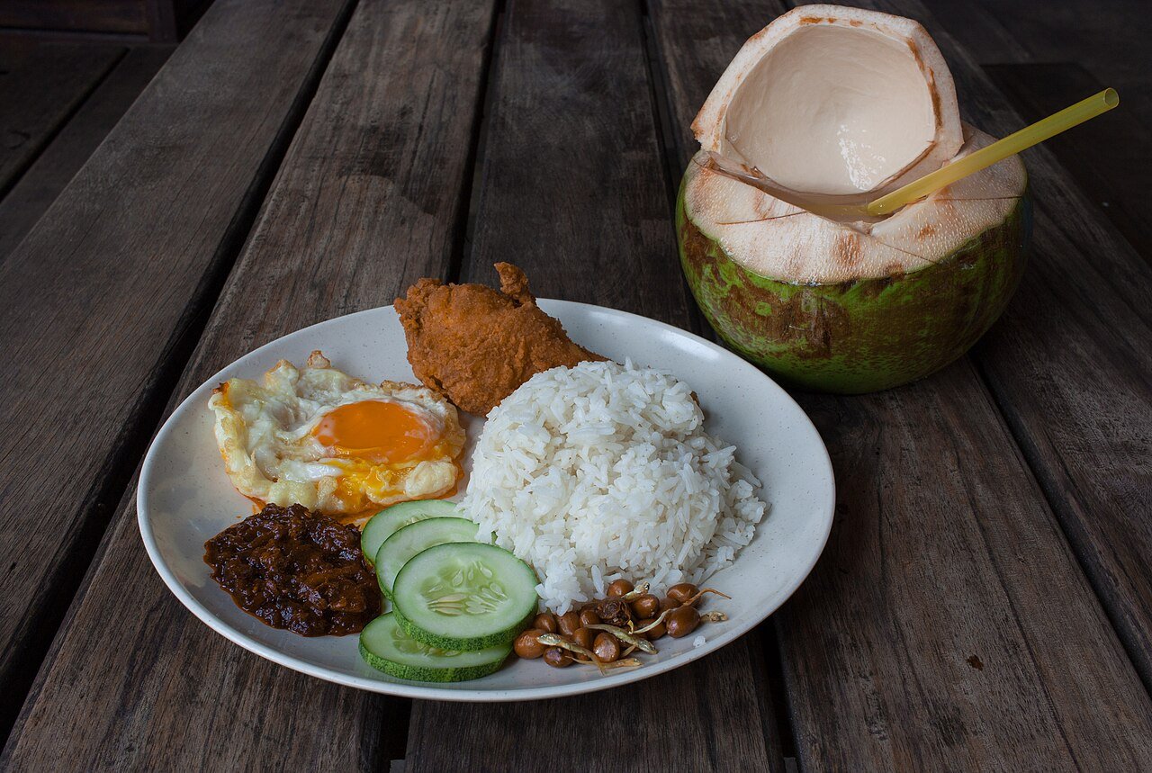 Nasi lemak with coconut rice, fried egg, sambal, cucumber, peanuts, and fried chicken, served with a young coconut