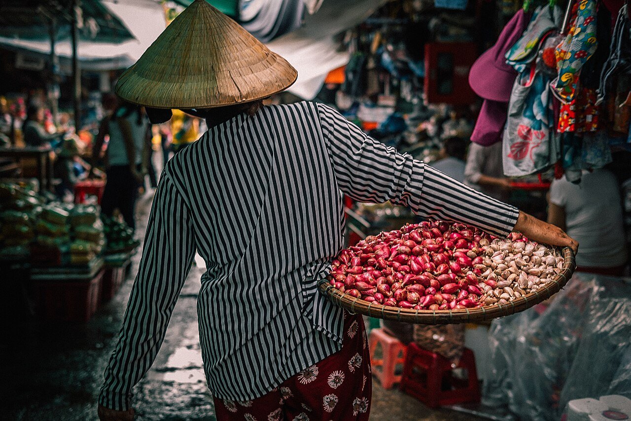 A vendor carrying a basket of shallots and garlic through Dong Ba market in Huế, Vietnam